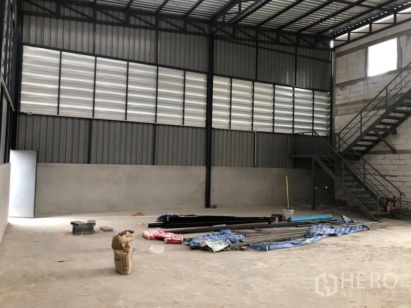 Warehouse for rent in Lam Luk Ka, Pathum Thani - Spacious warehouse interior with high steel truss roof, louvered panels, and metal staircase.