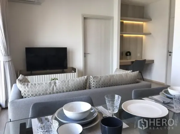 Condominium for rent in Huai Khwang, Bangkok - Dining setting with glass table facing the TV and a cozy study nook with shelves.