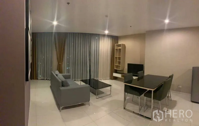 公寓 出租在Watthana, Bangkok - 夜景下的客厅，落地窗配长帘，沙发与餐桌，嵌灯照明。
