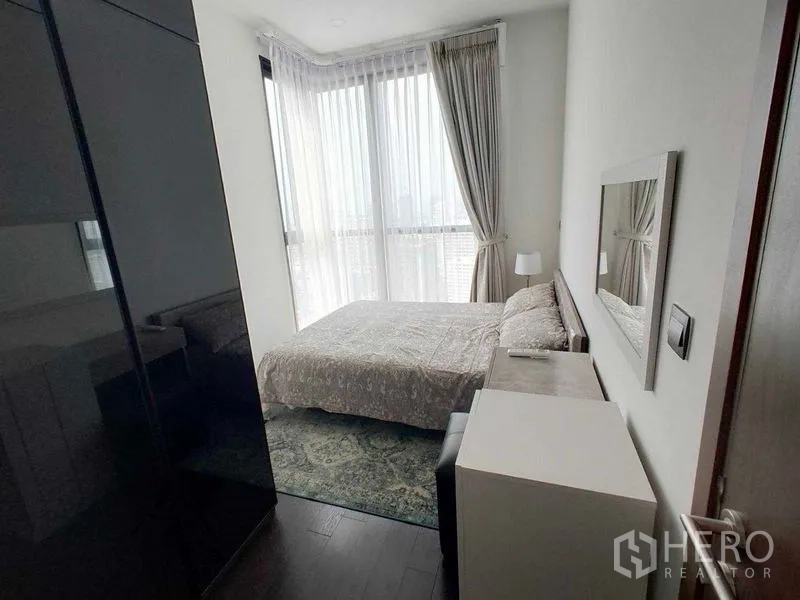 Condominium for rent in Watthana, Bangkok - Cozy bedroom with a bed, side table, and sheer curtains showing a city view.