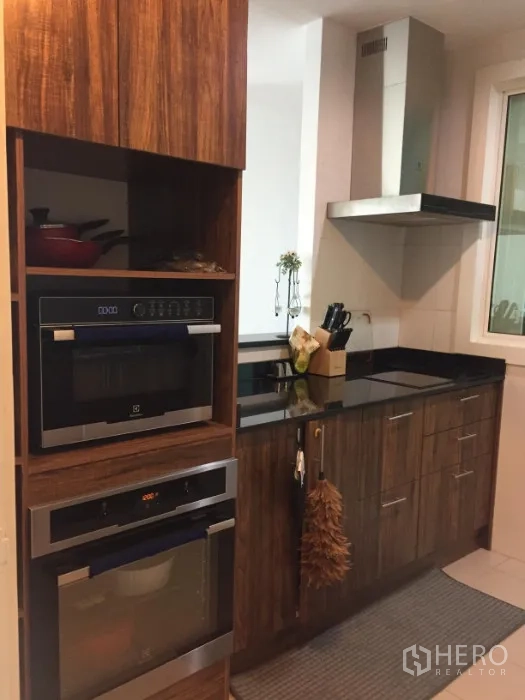 Condominium for rent or sale in Khlong Toei, Bangkok - Modern kitchen with wooden cabinetry, black granite countertops, built-in oven and microwave, and a range hood.