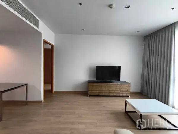Condominium for rent in Khlong Toei, Bangkok - Bright lounge with TV on a wood media unit, coffee table, and full-length curtains by a large window.