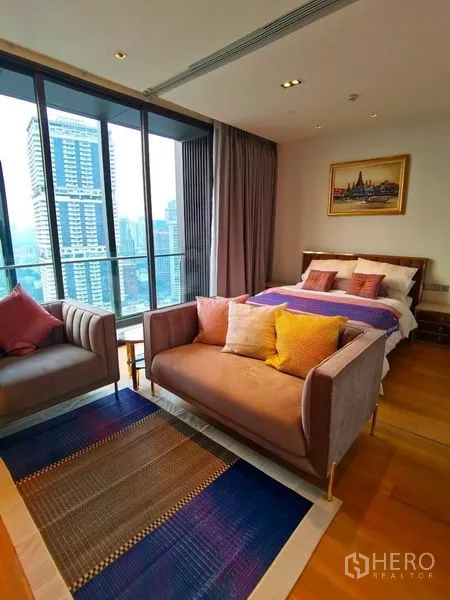 Condominium for rent in Khlong Toei, Bangkok - Bed and seating area beside wide windows overlooking a nearby high-rise.