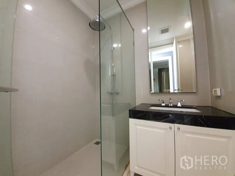 公寓 出租在Watthana, Bangkok - 卫浴间设玻璃淋浴房、顶喷花洒、长镜与黑色台面白色柜体。