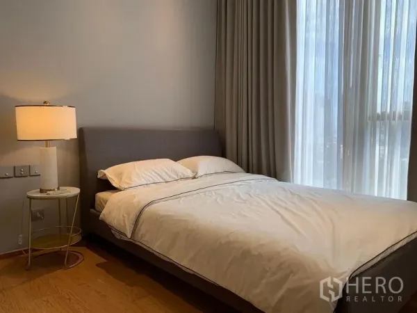 Condominium for rent in Khlong Toei, Bangkok - Cozy bedroom with upholstered bed, bedside lamp, tall curtains, and warm wood flooring.