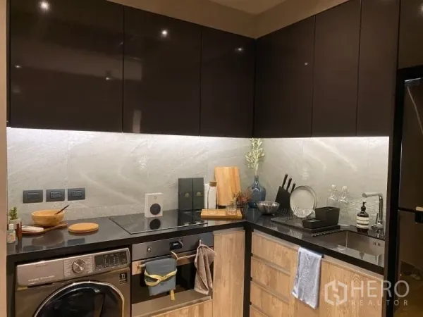Condominium for rent or sale in Watthana, Bangkok - L‑shaped kitchen with glossy cabinets, induction cooktop, oven, and washer‑dryer.