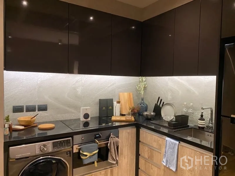 Condominium for rent or sale in Watthana, Bangkok - L‑shaped kitchen with glossy cabinets, induction cooktop, oven, and washer‑dryer.