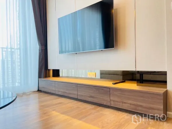 Condominium for rent in Khlong Toei, Bangkok - Close-up of a wall-mounted flat-screen TV over a long wooden console beside sheer balcony curtains.