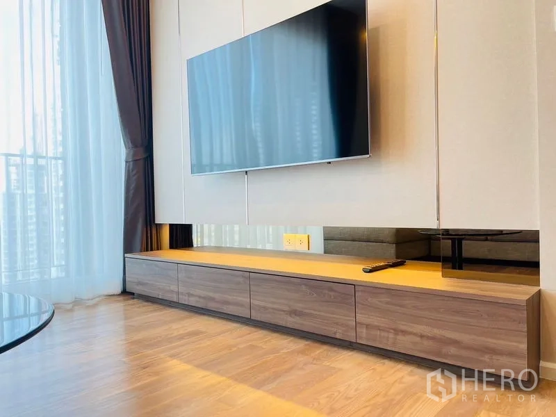 Condominium for rent in Khlong Toei, Bangkok - Close-up of a wall-mounted flat-screen TV over a long wooden console beside sheer balcony curtains.