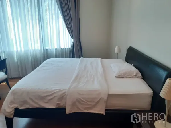 Condominium for rent in Khlong Toei, Bangkok - Primary bedroom with a large bed, bedside lamp, and wide curtained window offering natural light.
