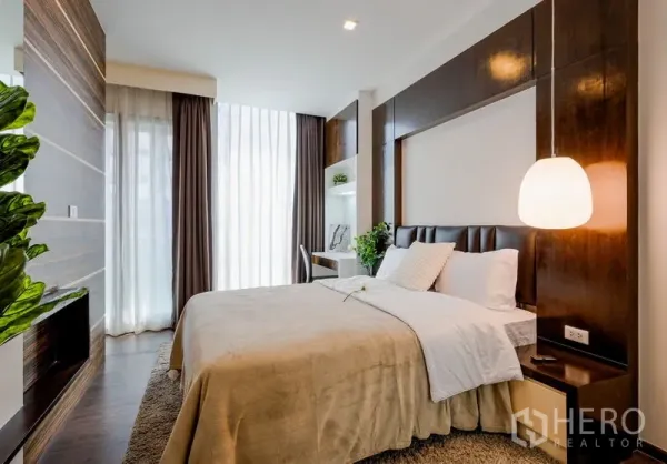 Condominium for sale in Khlong Toei, Bangkok - Bright master bedroom with large bed, wood paneling, pendant light, floor-to-ceiling curtains, and a small desk.