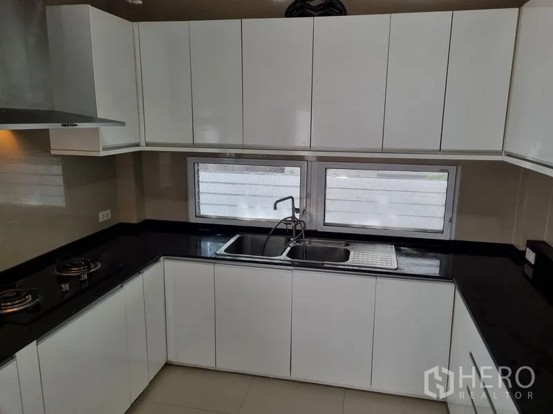 Detached House for sale in Chatuchak, Bangkok - U-shaped modern kitchen with white cabinetry, black countertops, gas hobs and a double sink beneath windows.