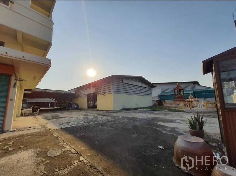 Warehouse for rent or sale in Mueang Pathum Thani, Pathum Thani - Spacious paved yard facing multiple warehouse bays at sunset with a small shrine nearby.