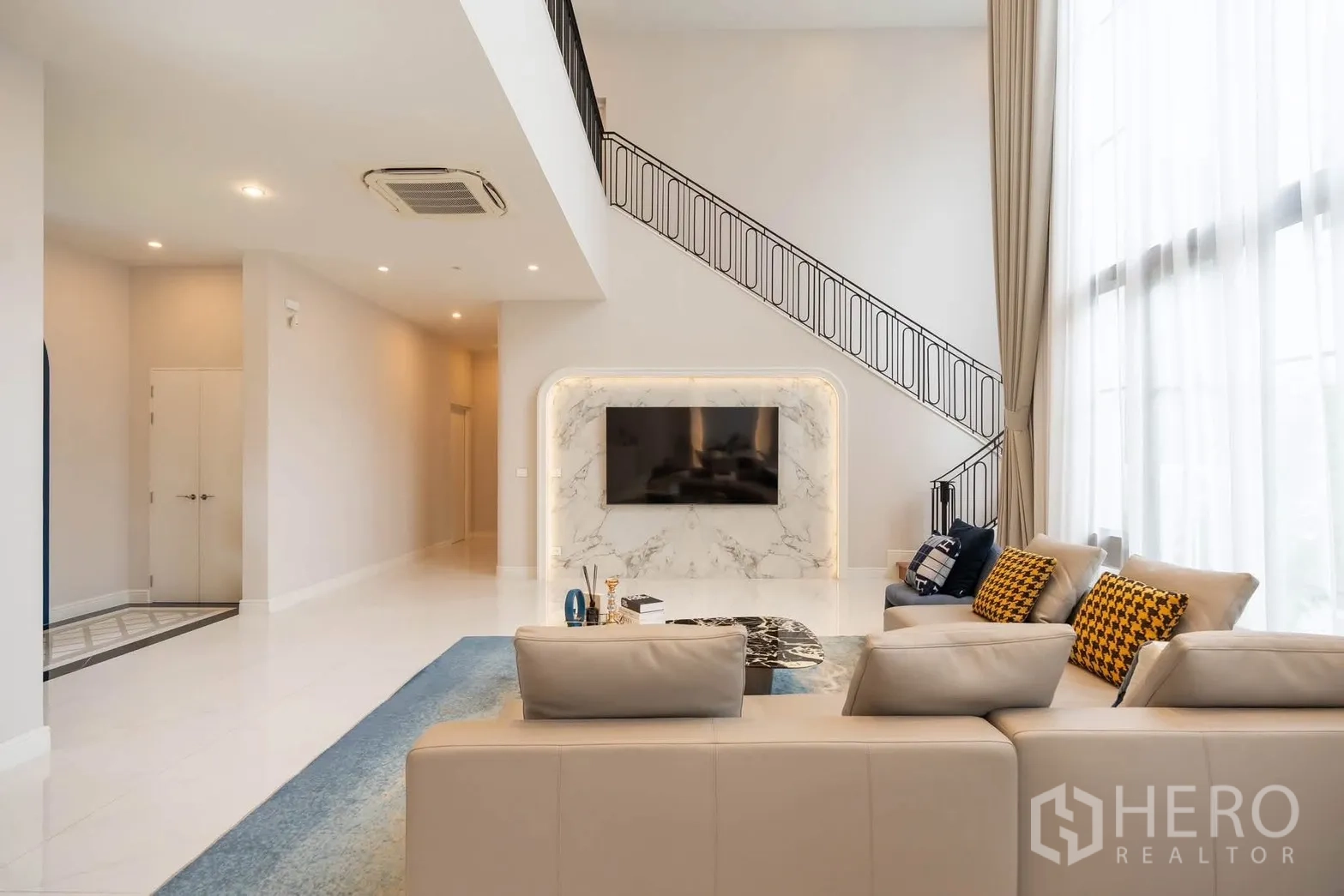 Detached House for rent or sale in Bang Kapi, Bangkok - Living room with marble TV wall, staircase with black railing, and tall curtains.