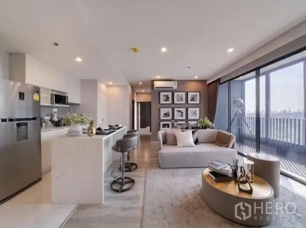 Condominium for rent in Bang Na, Bangkok - Sofa seating and kitchen facing full-height glass that opens to a balcony with city views.
