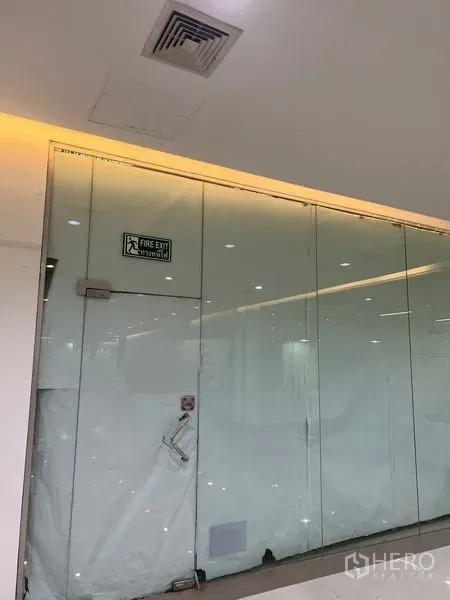 Retail Space for rent - Glass-fronted retail unit entrance with fire-exit sign and recessed lighting along a mall corridor in Watthana, Bangkok.