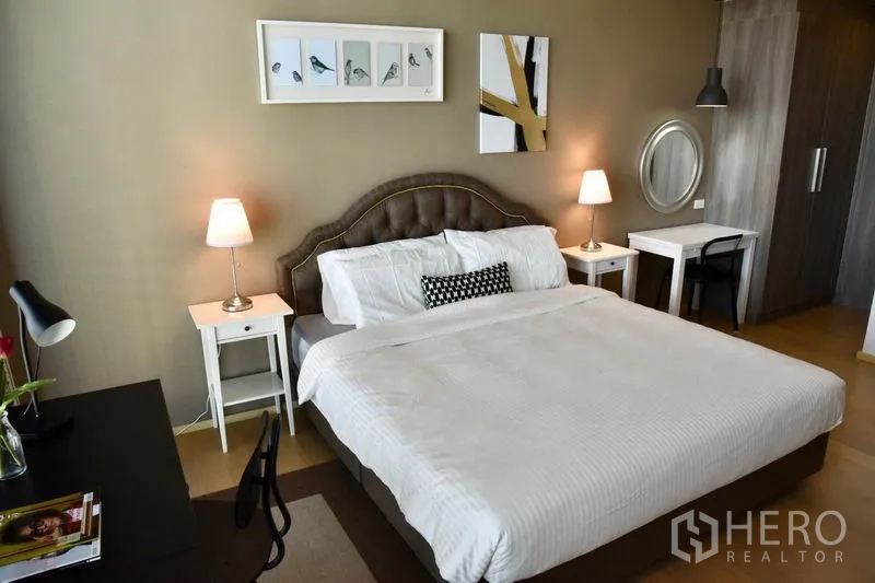 Condominium for rent in Phaya Thai, Bangkok - Bedroom with tufted headboard, artwork, bedside lamps, and built-in wardrobe beside a work desk.