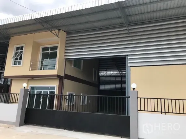 Warehouse for rent in Samut Sakhon - Warehouse exterior with wide roller shutter, two-storey office wing and gated entrance under a large roof canopy.