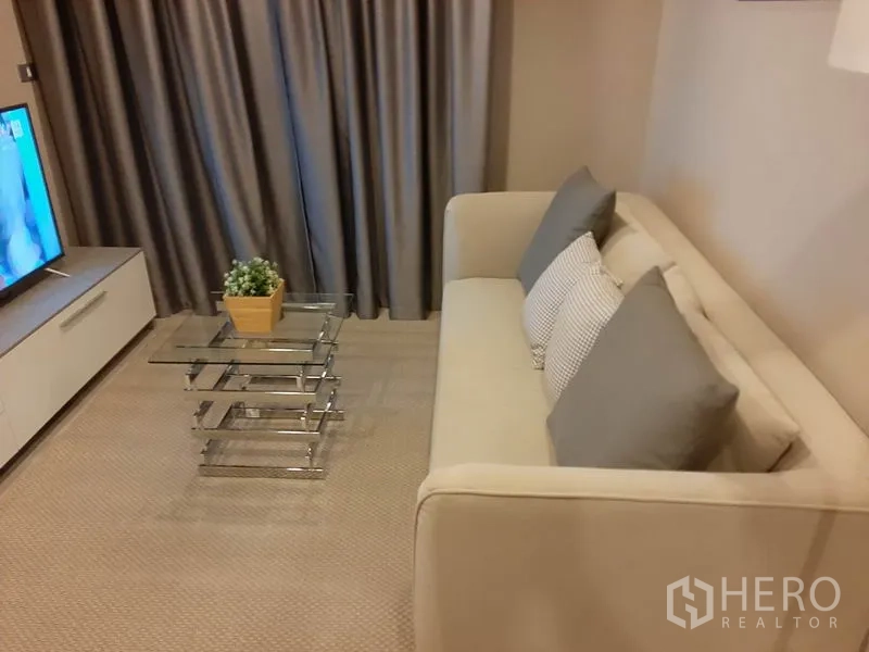 Condominium for rent or sale in Khlong Toei, Bangkok - Sofa and glass nesting coffee table beside the TV stand and curtains.