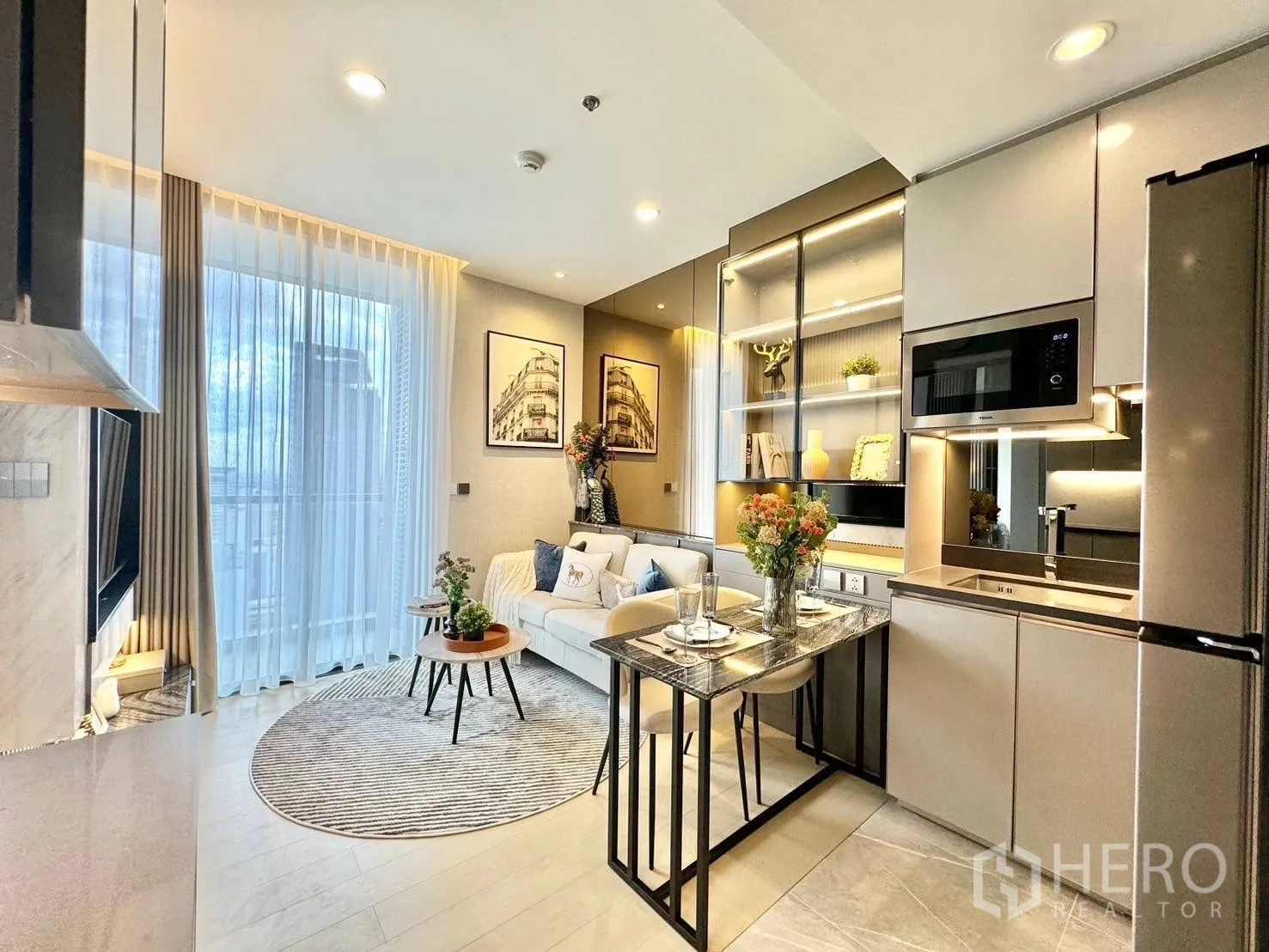 Condominium for rent in Ratchathewi, Bangkok - Bright living room with floor-to-ceiling window, round rug, and built-in shelves beside a sleek kitchenette.