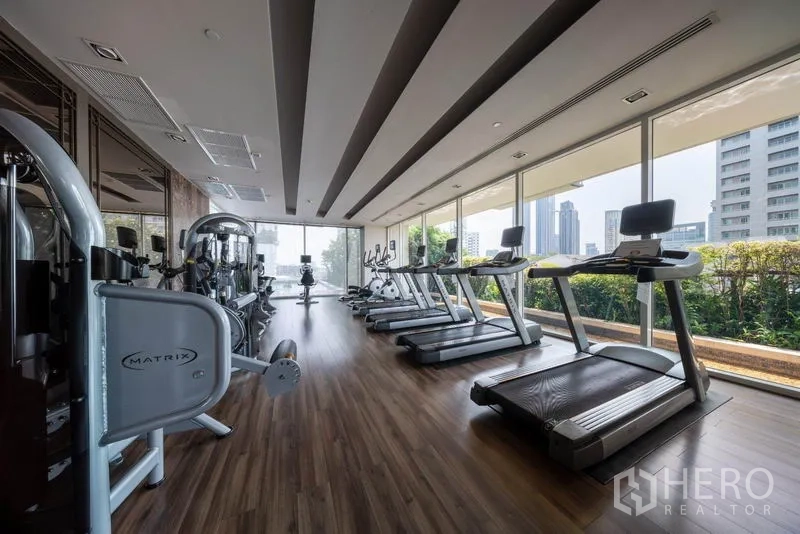 Condominium for rent in Watthana, Bangkok - Fully equipped gym with treadmills and weight machines facing floor-to-ceiling windows and city views.