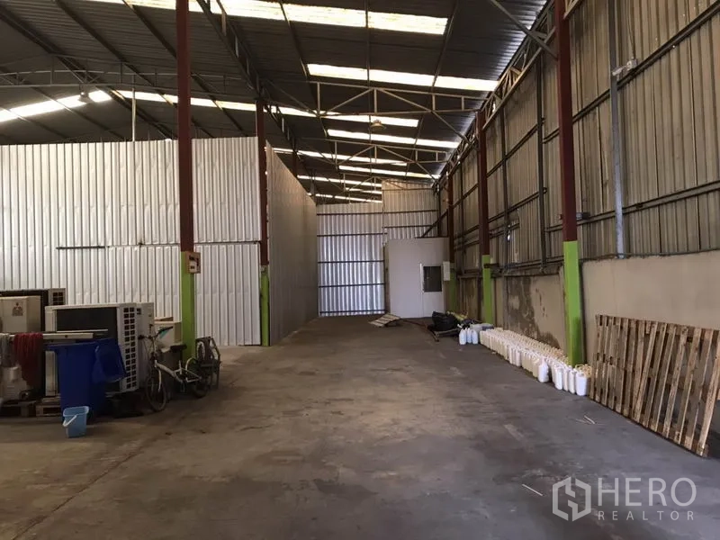 Warehouse for rent in Bang Na, Bangkok - Long aisle of the warehouse showing metal partitions, a small utility room and exposed roof trusses over a concrete floor.
