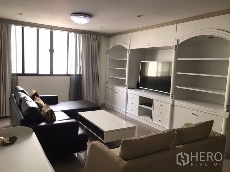 公寓 出租在Watthana, Bangkok - 明亮客厅，白色定制电视墙与开放式书架，黑色皮沙发与茶几，大窗配窗帘。