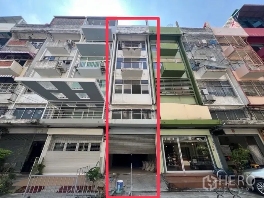 อาคารพาณิชย์ ให้เช่าในBangkok - ด้านหน้าอาคารพาณิชย์ 4 ชั้น มีระเบียงและประตูม้วน โดดเด่นด้วยกรอบสีแดง ตั้งอยู่ท่ามกลางตึกแถว.