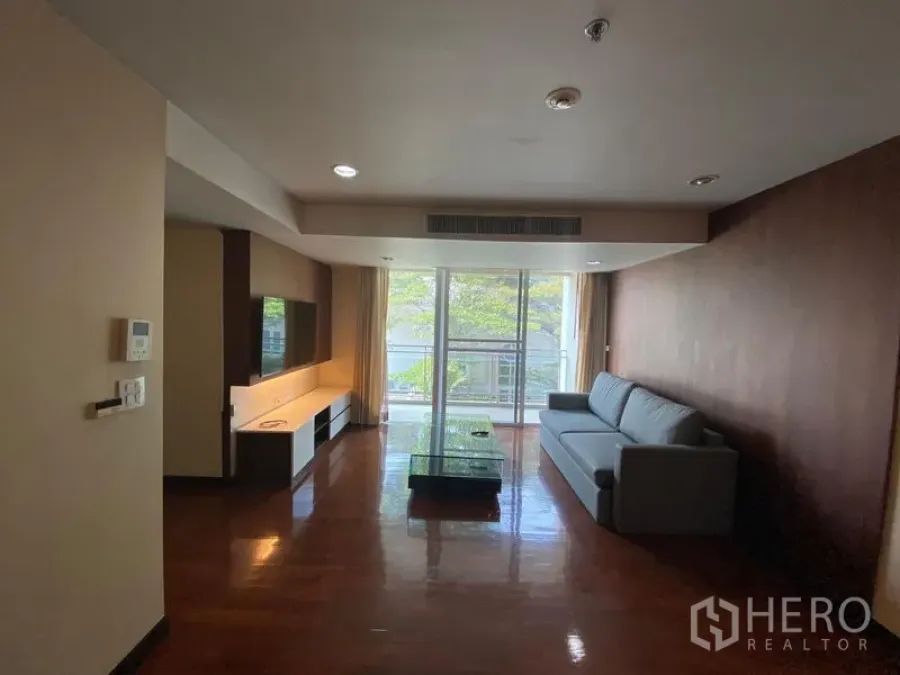 公寓 出租在Watthana, Bangkok - 客厅宽敞，配沙发、电视墙、木地板与通向绿景阳台的推拉玻璃门。