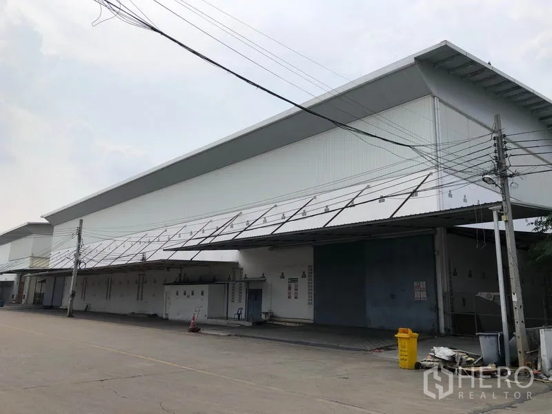 Factory for rent in Bang Bua Thong, Nonthaburi - Wide exterior of a modern white factory with long covered loading bays along a paved road.