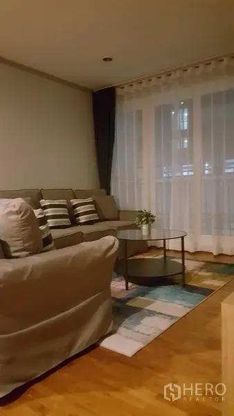 Condominium for rent in Watthana, Bangkok - Evening view of the living area with floor-to-ceiling sheer curtains, gray sectional sofa, and round coffee table.