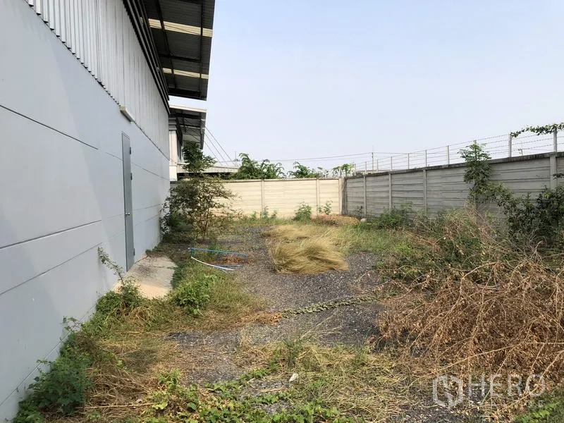 Warehouse for rent or sale in Lam Luk Ka, Pathum Thani - Fenced side yard with gravel and greenery running along the warehouse exterior.