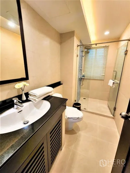 Condominium for rent in Bang Rak, Bangkok - Modern bathroom with glass-enclosed shower, vanity, and beige tiles.