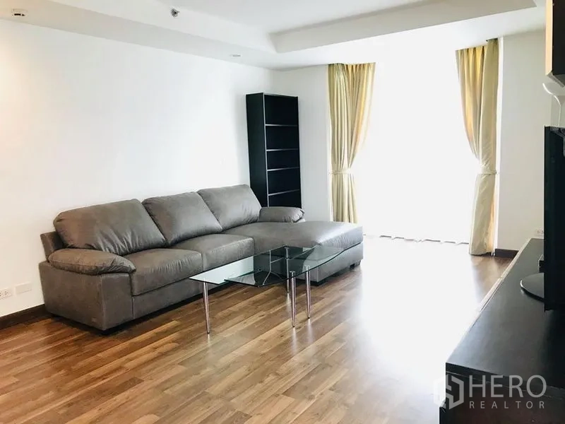 公寓 出租在Pathum Wan, Bangkok - 宽敞起居区，灰色转角沙发与玻璃茶几，高窗配窗帘。