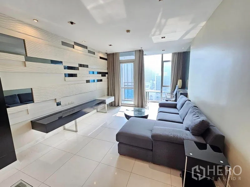คอนโด ให้เช่าหรือขายในPathum Wan, Bangkok - ห้องนั่งเล่นสว่าง โซฟา L‑shape ผนังตกแต่ง และหน้าต่างเต็มบานเปิดสู่ระเบียงชมวิวเมือง