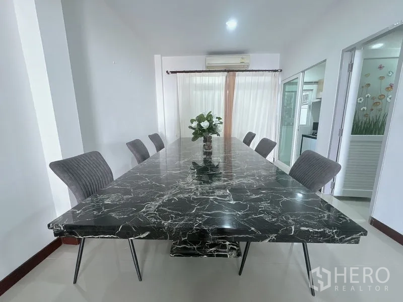 联排别墅 出租在Prawet, Bangkok - 明亮餐厅，长条黑色大理石餐桌、灰色软椅与薄纱窗帘。