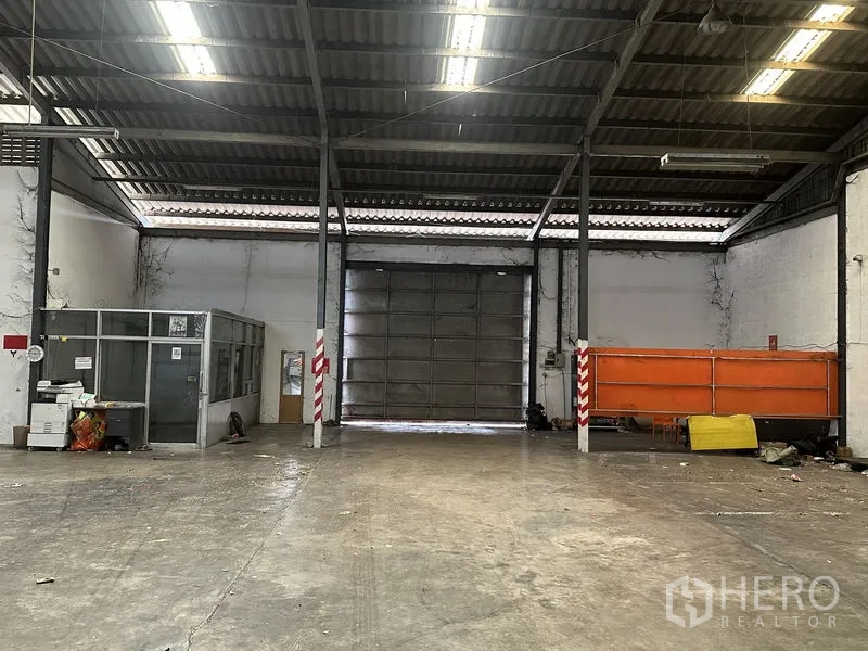 Warehouse for rent in Khan Na Yao, Bangkok - Rear interior view showing a large roller door, small glass office and storage area.