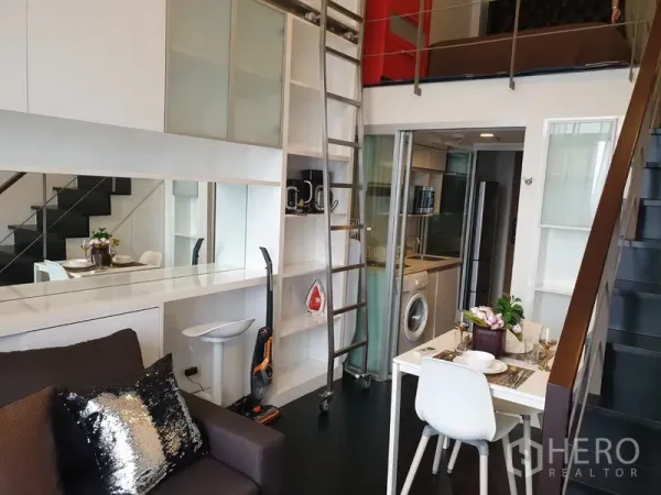 Condominium for rent in Khlong Toei, Bangkok - Loft-style living area with built-in shelves, rolling ladder, compact glass-enclosed kitchen, and a small dining set.