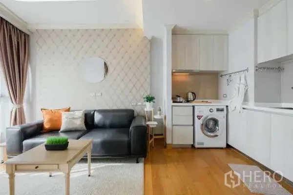 Condominium for rent in Khlong Toei, Bangkok - Open-plan living with black leather sofa, compact white kitchen and in-unit washing machine.