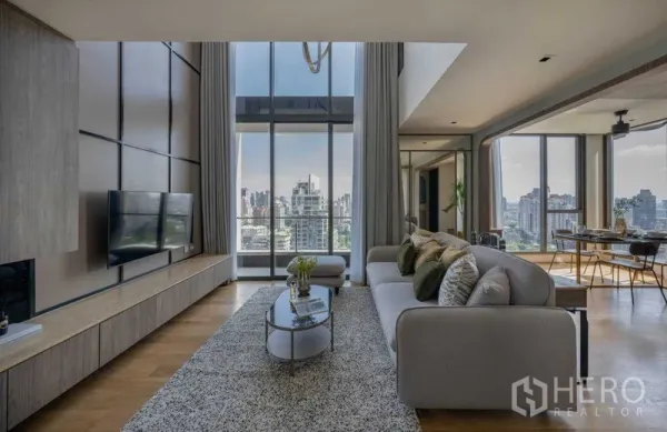 Condominium for sale in Khlong Toei, Bangkok - Spacious living area with sofa and rug facing a panoramic city view through tall glass walls.