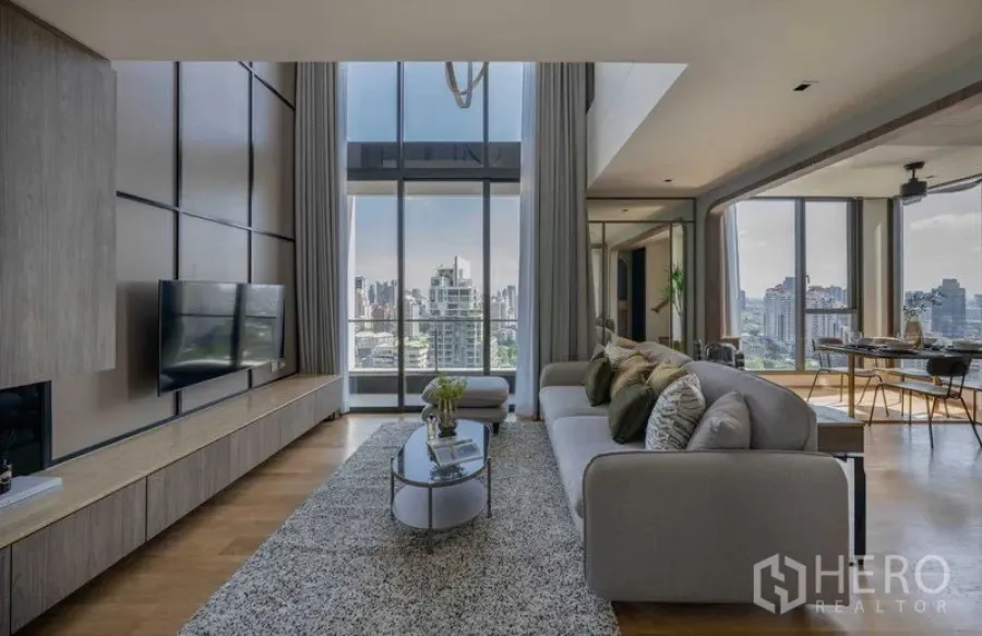 Condominium for sale in Khlong Toei, Bangkok - Spacious living area with sofa and rug facing a panoramic city view through tall glass walls.