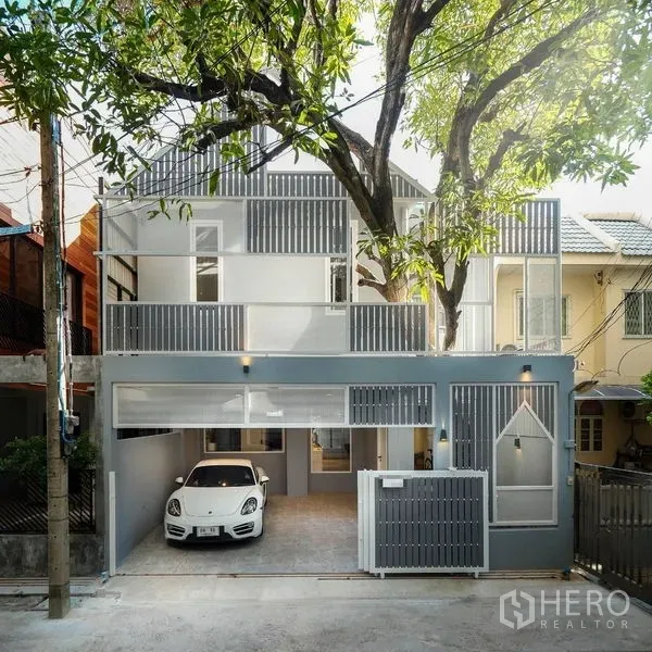 Townhouse for rent in Watthana, Bangkok - Modern townhouse exterior with gray facade, balconies, sliding gate and carport shaded by a tree.