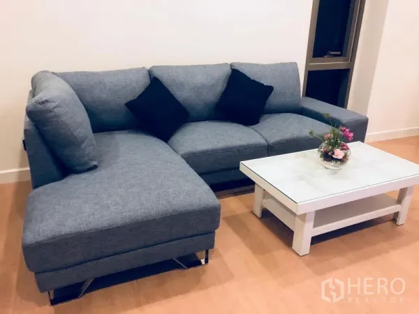 Condominium for rent or sale in Bang Rak, Bangkok - Living room corner with a grey L‑shaped sofa, black cushions and a white glass‑top coffee table on wood flooring.
