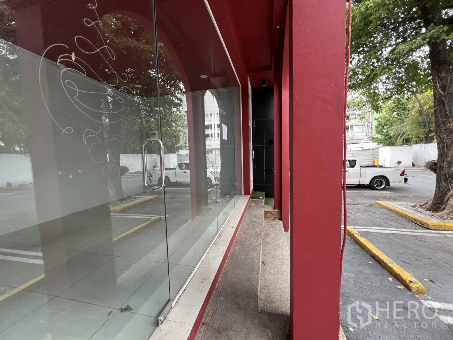 พื้นที่ขายของ for rent in Watthana, Bangkok - ระเบียงกระจกหน้าร้านพร้อมเสาแดงและมือจับ มองเห็นลานจอดรถและต้นไม้