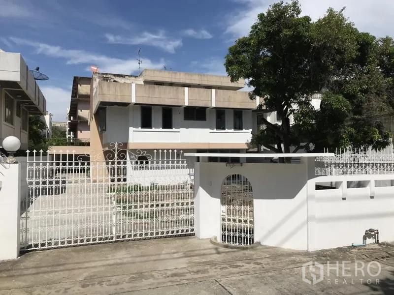 独立式住宅 出租或出售在Khlong Toei, Bangkok - 从前门看到的两层白色房屋，金属围栏与宽车道，绿树环绕，靠近BTS Phra Khanong。