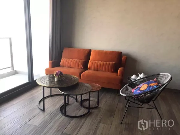 Condominium for sale in Watthana, Bangkok - Cozy lounge corner with an orange sofa, nested round metal coffee tables, and a large glass door to the balcony.