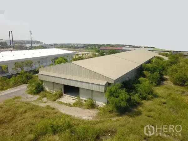 Factory for sale in Samut Prakan - Aerial view of a long metal-roof factory warehouse surrounded by greenery in an industrial area.