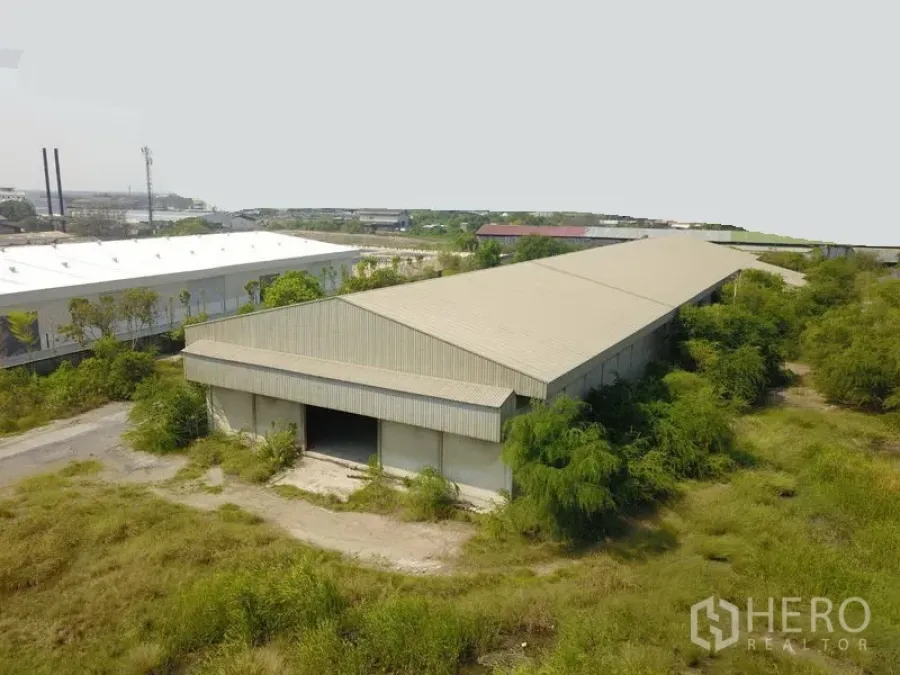 Factory for sale in Samut Prakan - Aerial view of a long metal-roof factory warehouse surrounded by greenery in an industrial area.