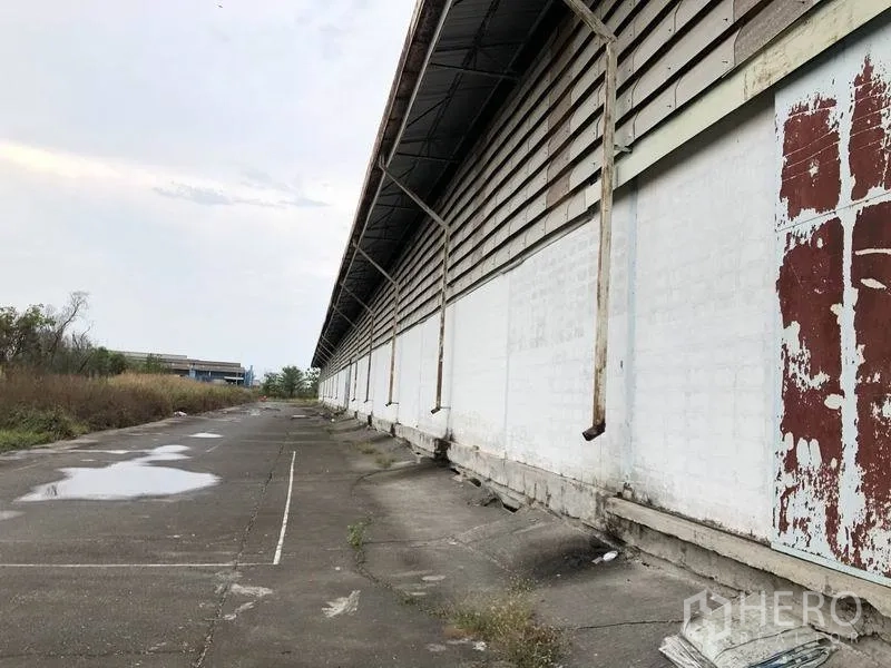 Warehouse for rent in Samut Prakan - Exterior side of the warehouse with long white wall, driveway for trucks, and drainage.