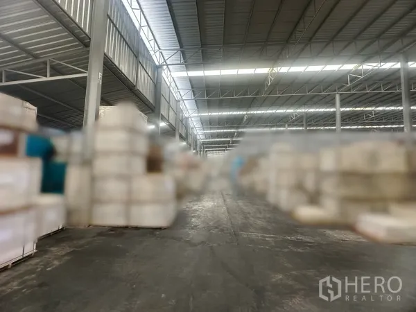 Warehouse for rent in Bang Yai, Nonthaburi - Aisle between stored goods in a metal-frame warehouse with continuous skylights and concrete flooring.